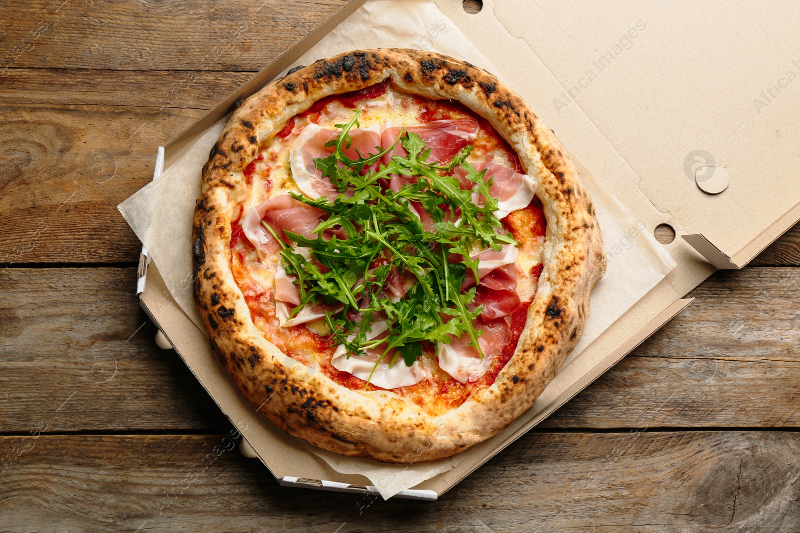 Photo of Tasty pizza with meat and arugula in cardboard box on wooden table, top view