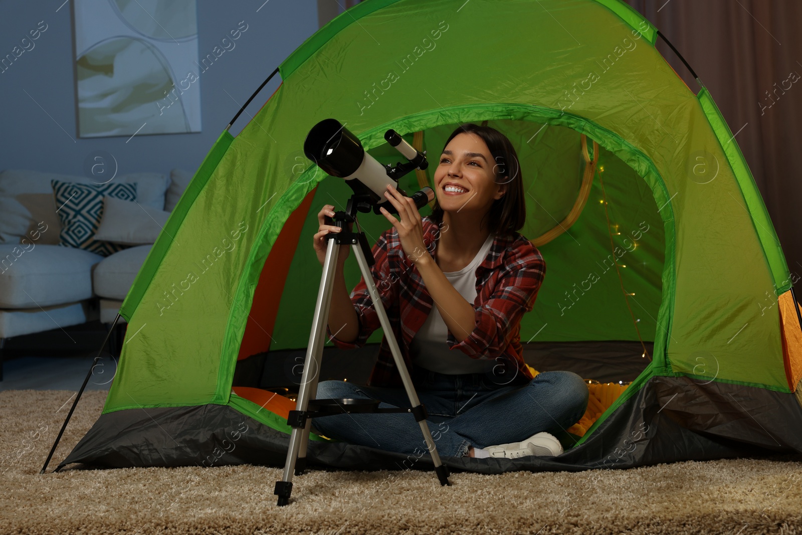 Photo of Young woman using telescope to look at stars while sitting in camping tent indoors