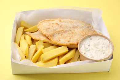 Photo of Delicious fish and chips with tasty sauce in paper box on yellow table, closeup
