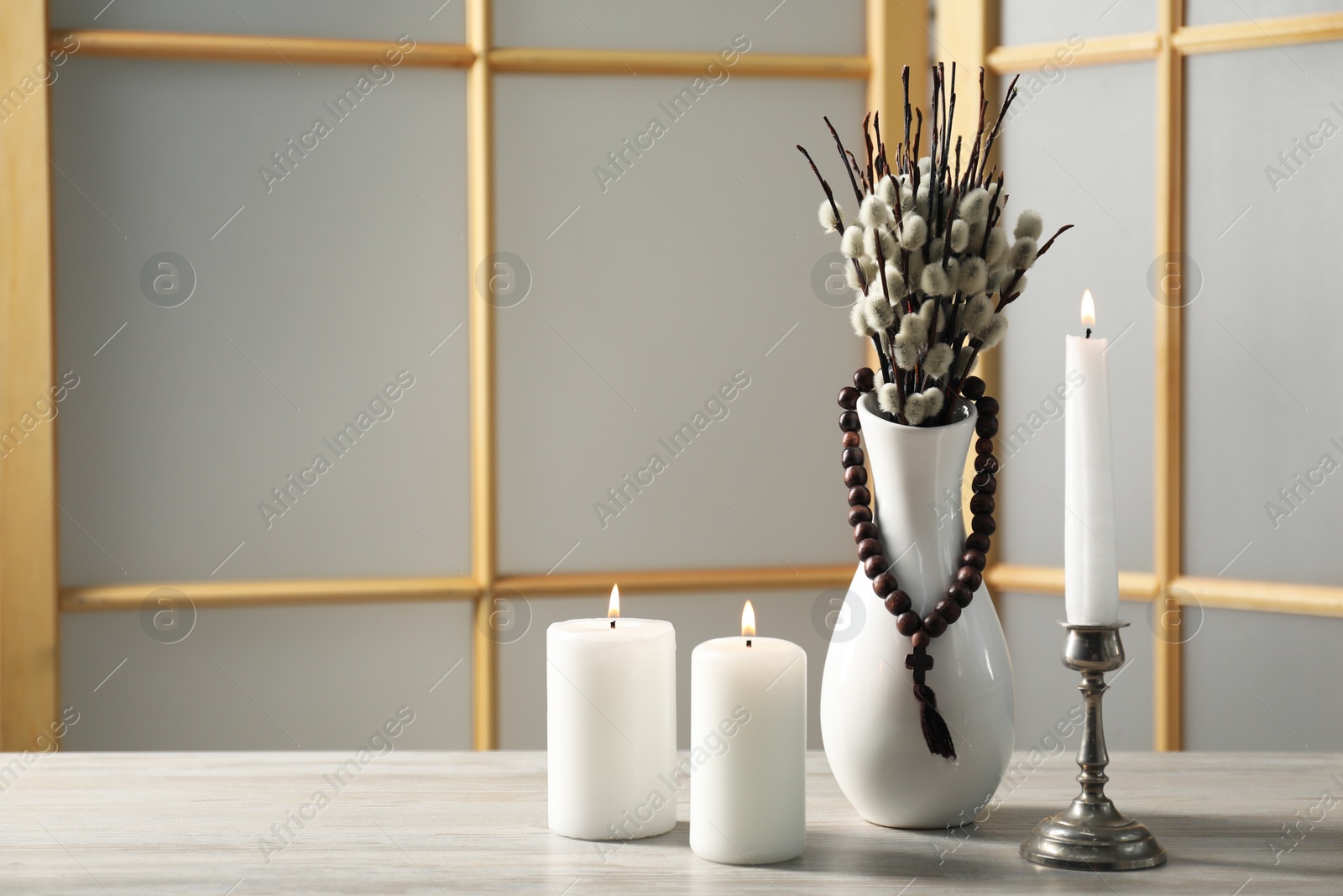 Photo of Rosary beads, burning candles and vase of willow branches on wooden table, space for text