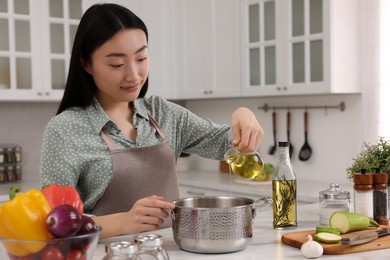 Photo of Cooking process. Beautiful woman pouring oil from bottle into pot at white countertop in kitchen