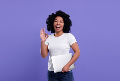 Happy young woman with laptop showing ok gesture on purple background