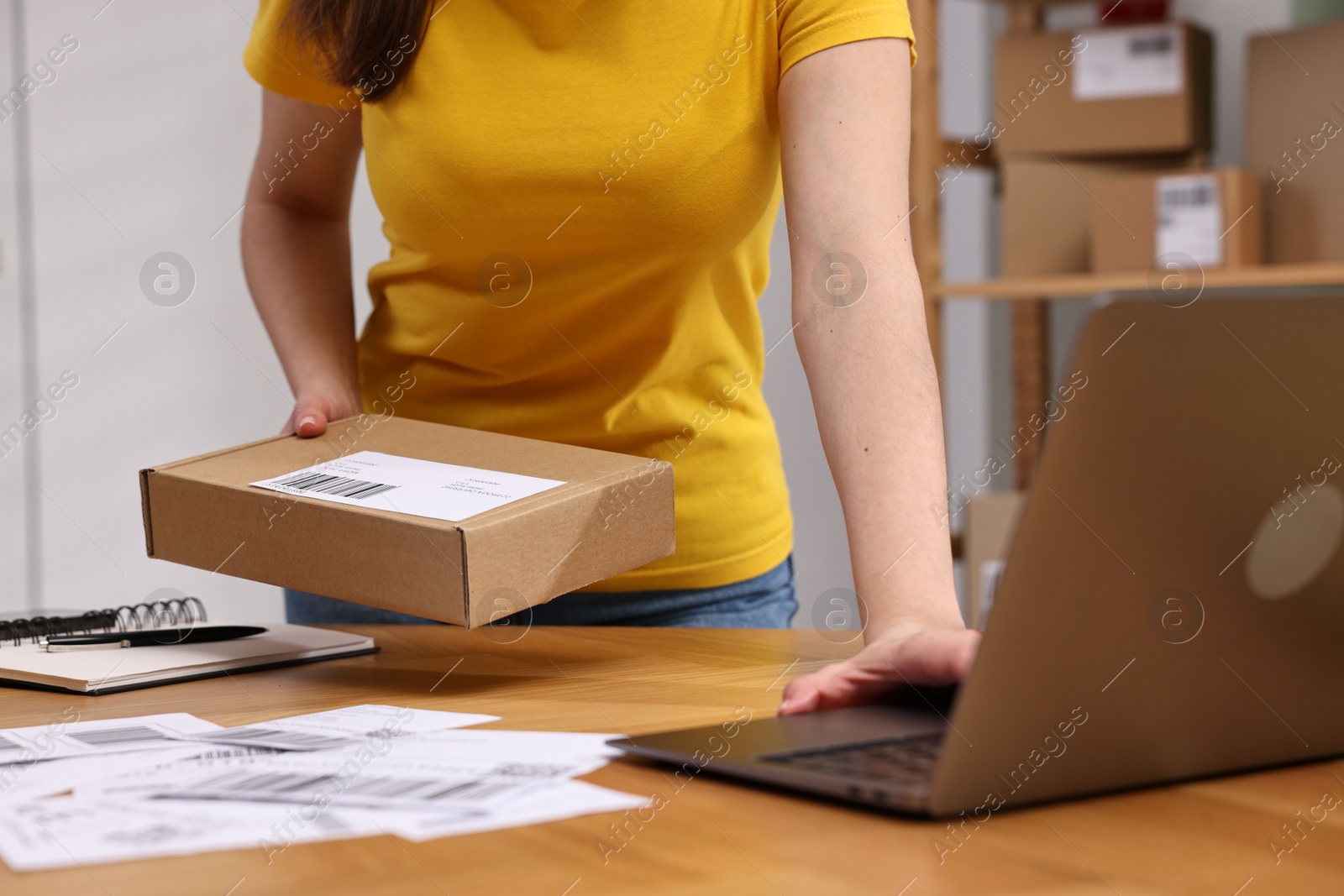 Photo of Parcel packing. Post office worker with box using laptop at wooden table indoors, closeup