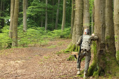 Man with hunting rifle and backpack wearing camouflage in forest, back view. Space for text