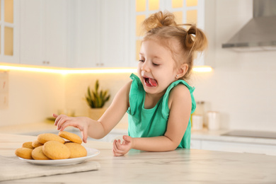 Photo of Cute little girl at table with cookies in kitchen