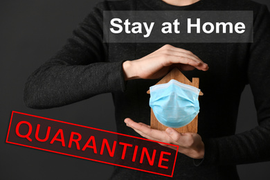 Image of Stay at home during coronavirus quarantine. Woman holding wooden house model with medical mask on dark background
