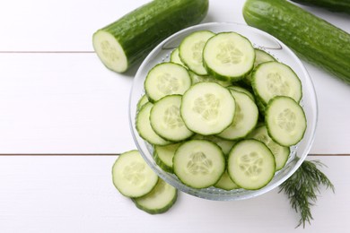 Photo of Cut cucumber in glass bowl, fresh vegetables and dill on white wooden table, flat lay. Space for text