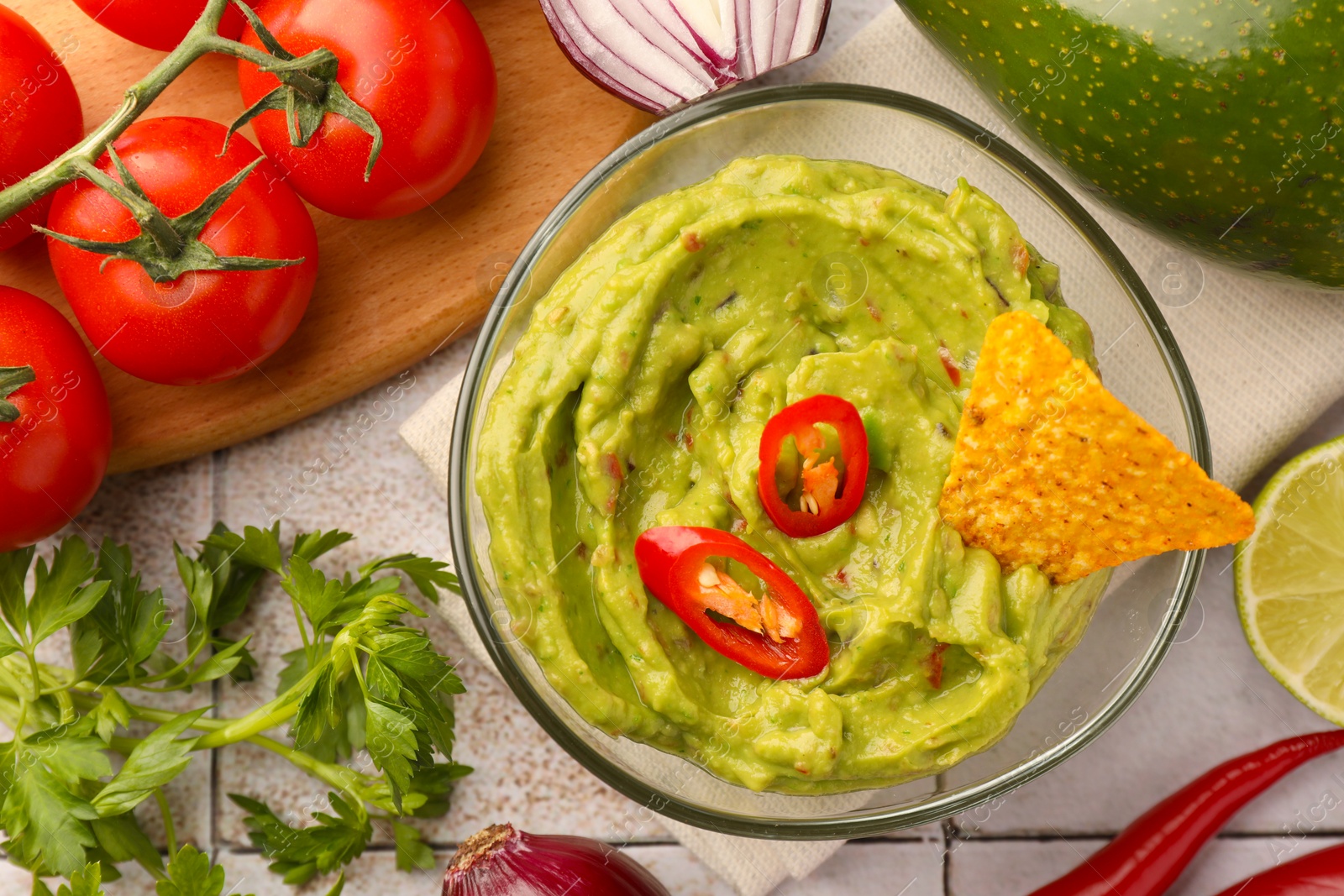 Photo of Bowl of delicious guacamole, nachos chip and ingredients on white tiled table, flat lay