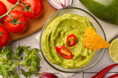 Bowl of delicious guacamole, nachos chip and ingredients on white tiled table, flat lay