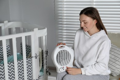 Photo of Young woman adjusting temperature on modern electric fan heater in child room