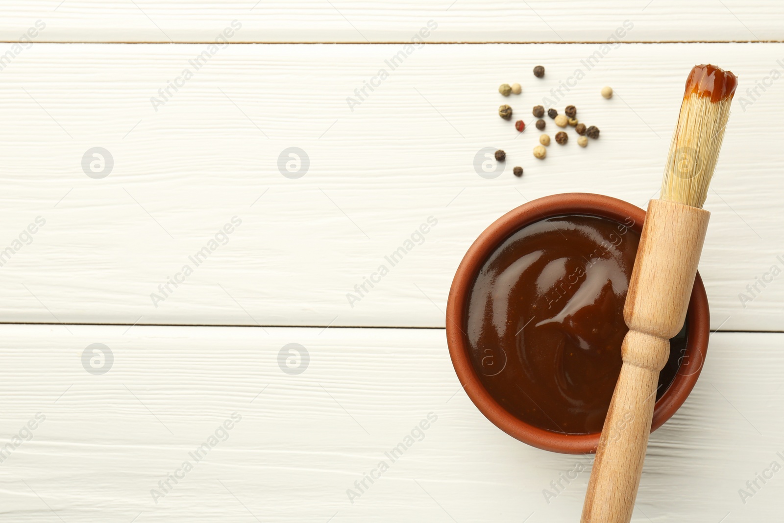 Photo of Tasty barbeque sauce in bowl, brush and peppercorns on white wooden table, top view. Space for text