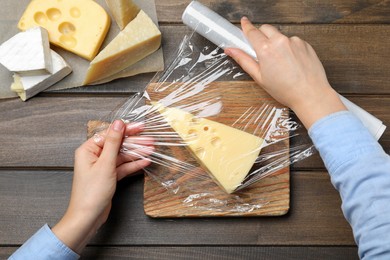 Photo of Woman putting plastic food wrap over block of cheese at wooden table, top view
