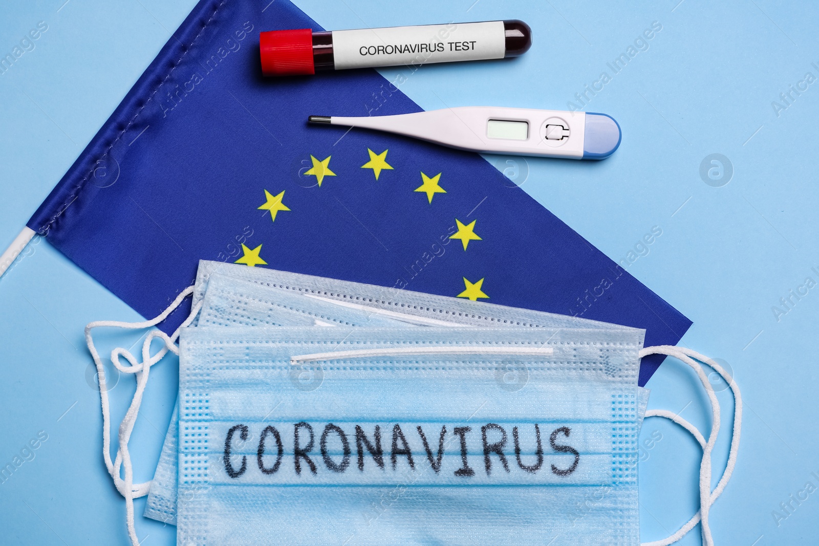 Photo of European Union flag, protective masks, thermometer and test tube with blood sample on light blue background, flat lay. Coronavirus outbreak