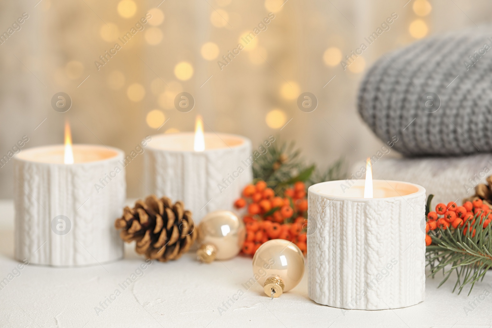 Photo of Holders with burning candles and decoration on white table against blurred Christmas lights