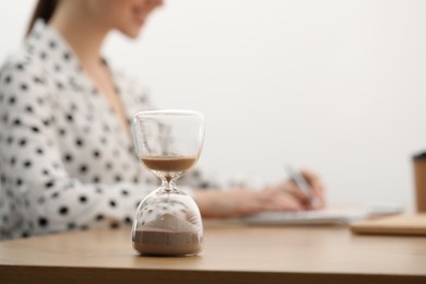 Hourglass with flowing sand on desk. Woman taking notes while using calculator indoors, selective focus