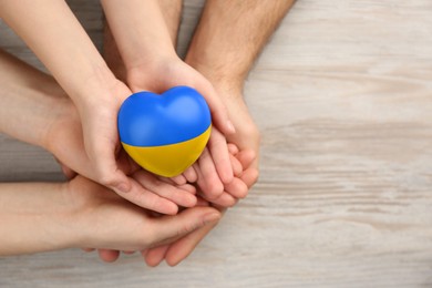 Image of Stop war in Ukraine. Family holding heart shaped toy with colors of Ukrainian flag in hands at wooden table, top view