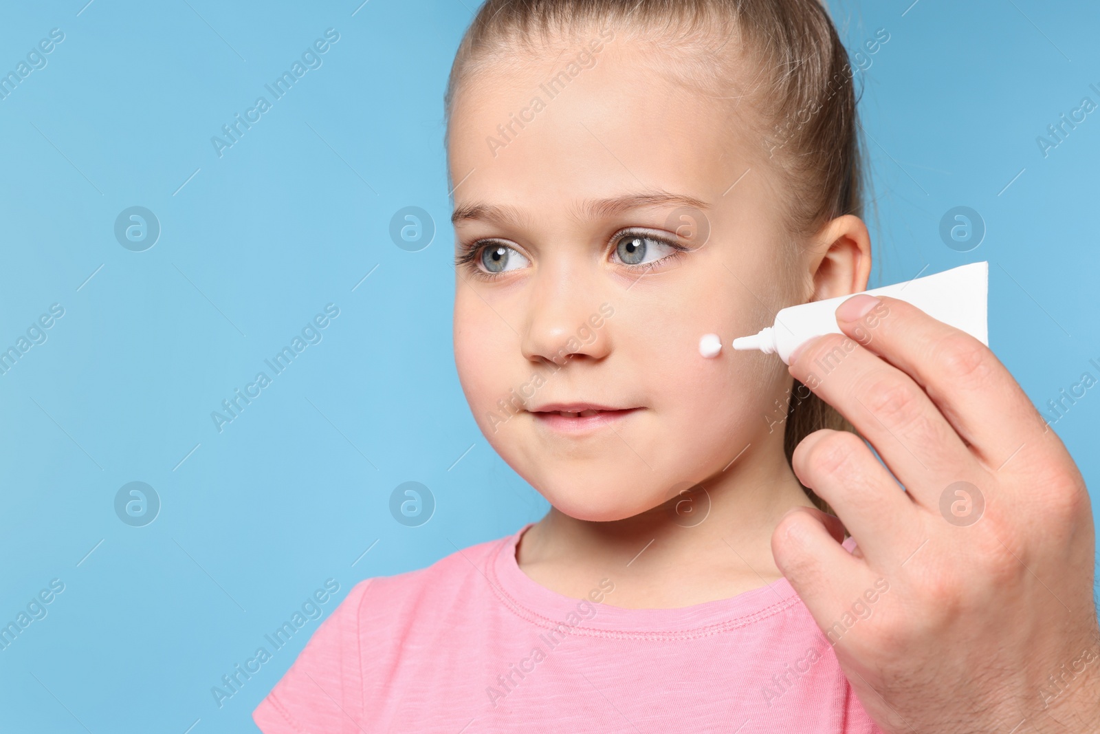 Photo of Father applying ointment onto his daughter's cheek on light blue background, space for text