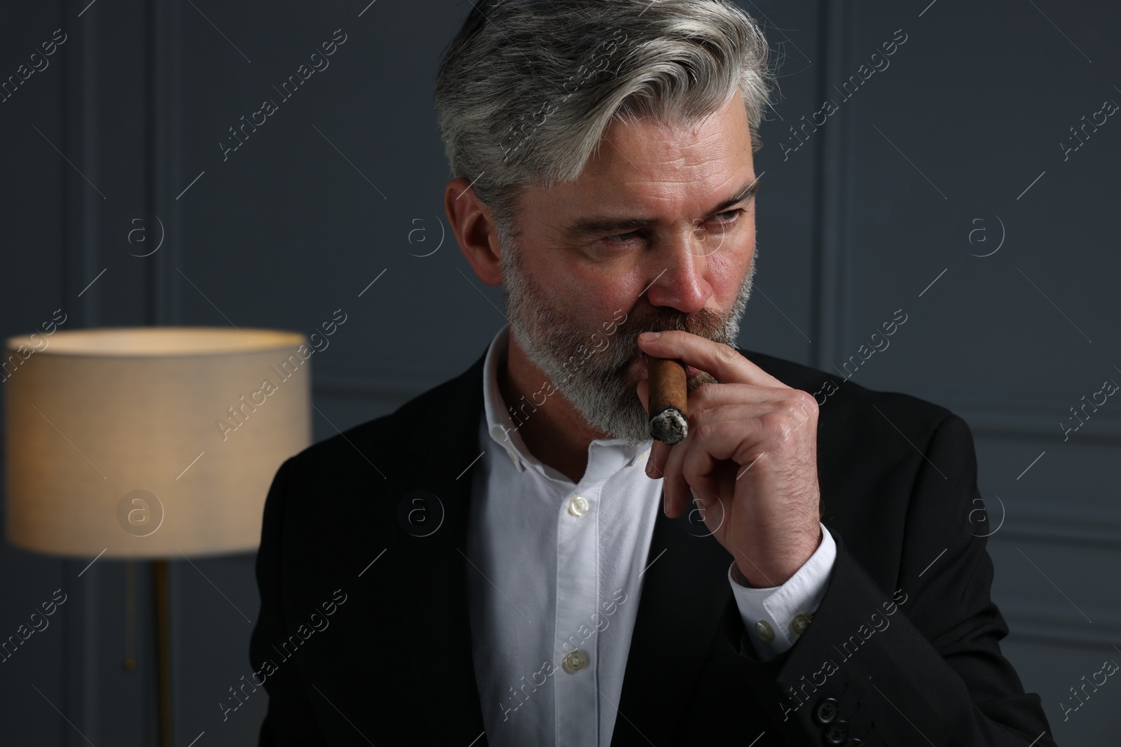 Photo of Handsome bearded man smoking cigar near dark grey wall indoors