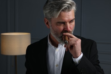 Handsome bearded man smoking cigar near dark grey wall indoors
