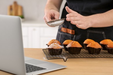 Photo of Man decorating muffins with powdered sugar while watching online cooking course via laptop in kitchen, closeup. Time for hobby