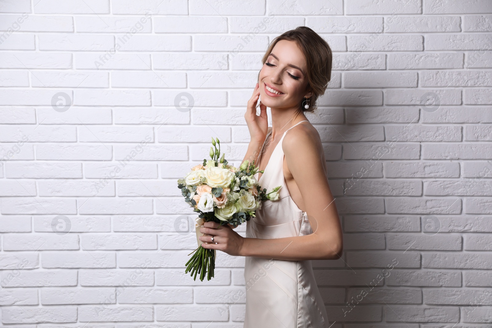 Photo of Young bride with elegant hairstyle holding wedding bouquet near white brick wall