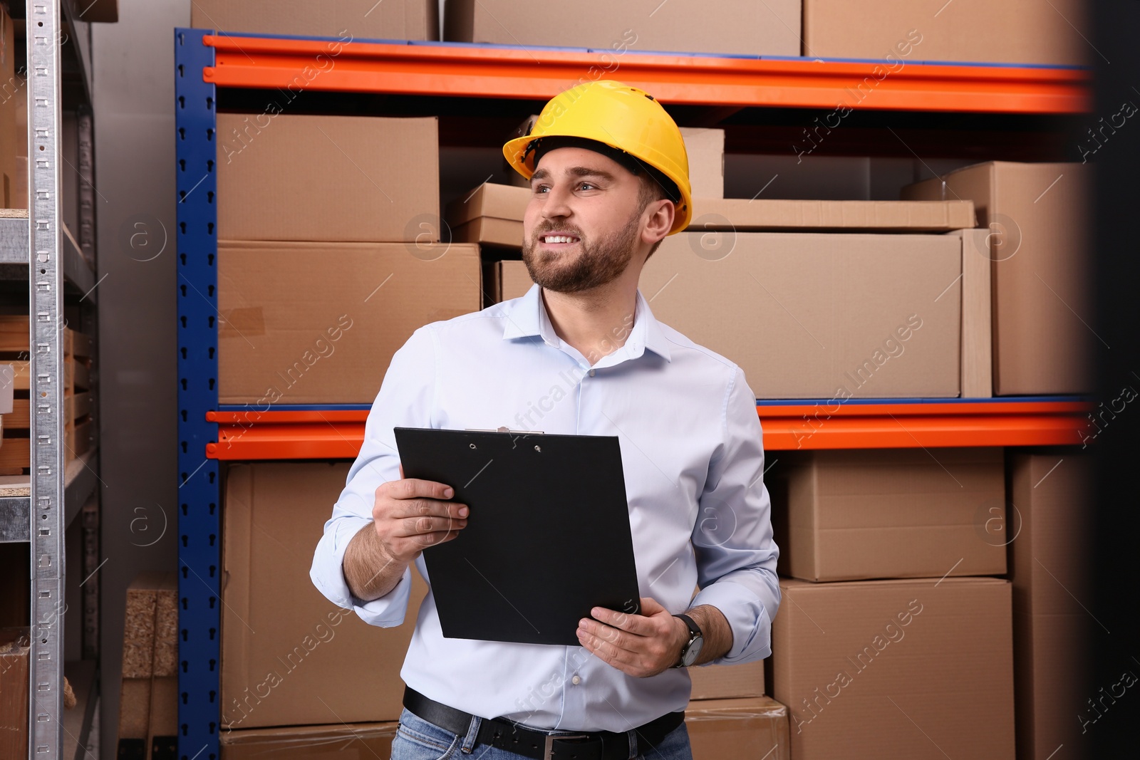 Photo of Young man with clipboard near rack of cardboard boxes at warehouse