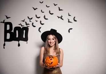Woman in witch hat with jack o'lantern posing near white wall decorated for Halloween