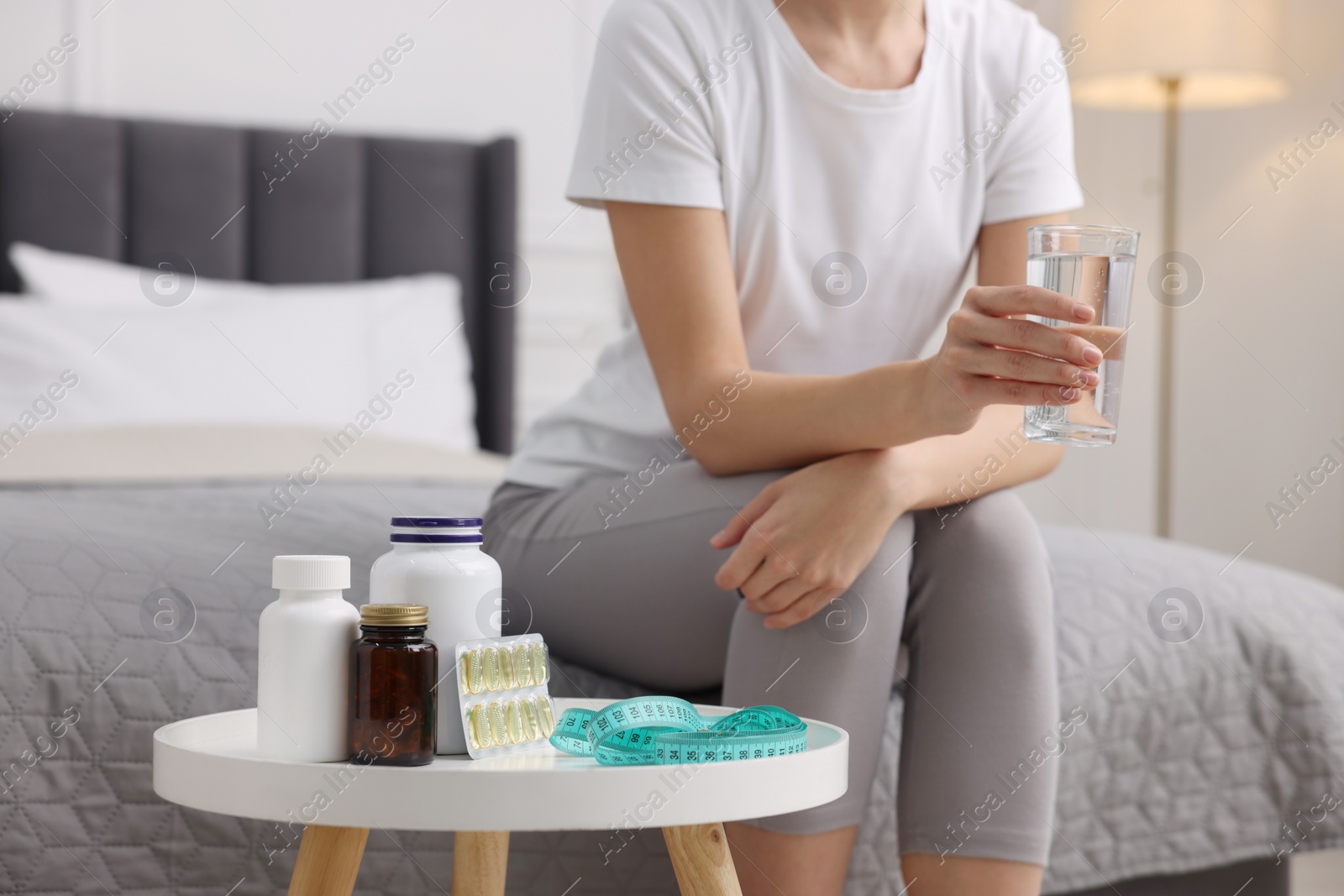 Photo of Woman holding glass of water near table with pills and measuring tape in room, closeup. Weight loss