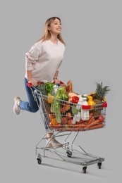 Young woman with shopping cart full of groceries on grey background