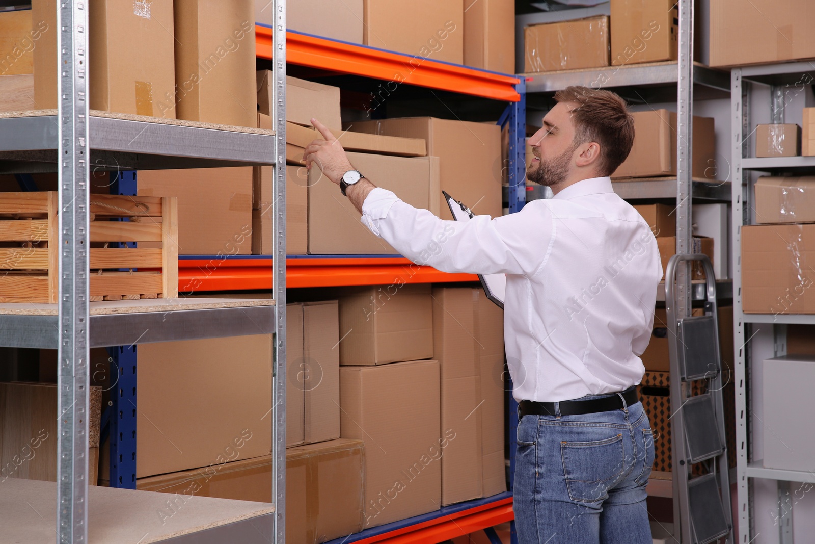 Photo of Young businessman with clipboard near rack of cardboard boxes at warehouse