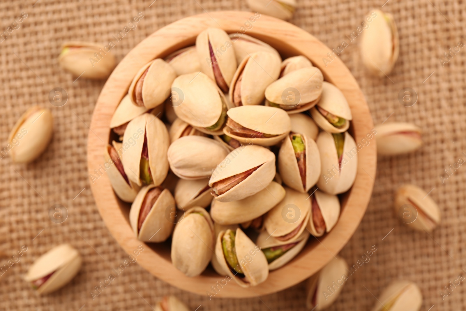 Photo of Tasty pistachios in bowl on burlap fabric, top view