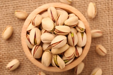 Photo of Tasty pistachios in bowl on burlap fabric, top view