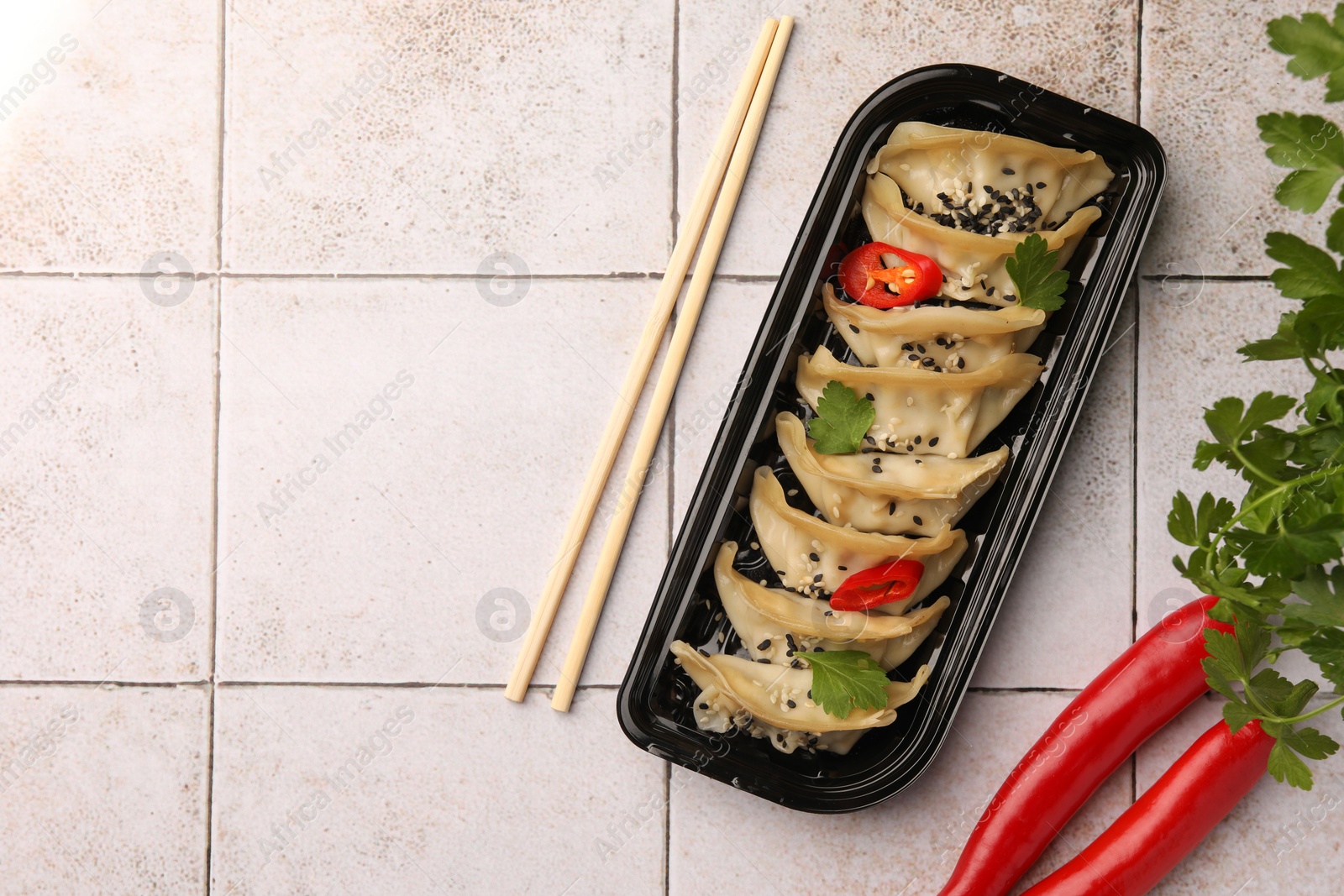 Photo of Delicious gyoza (asian dumplings) with chili peppers, parsley and chopsticks on light tiled table, flat lay. Space for text