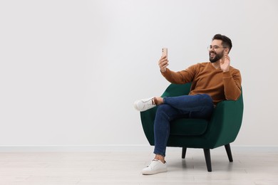 Photo of Handsome man having video chat via smartphone while sitting in armchair near white wall indoors, space for text