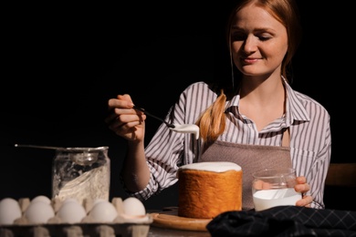 Photo of Young woman decorating traditional Easter cake with glaze at table against black background