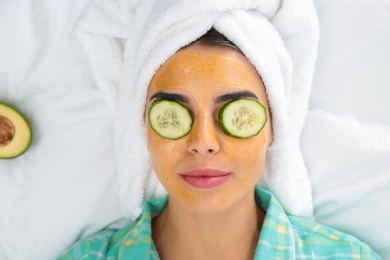 Photo of Young woman with facial mask and cucumber slices lying on bed, top view
