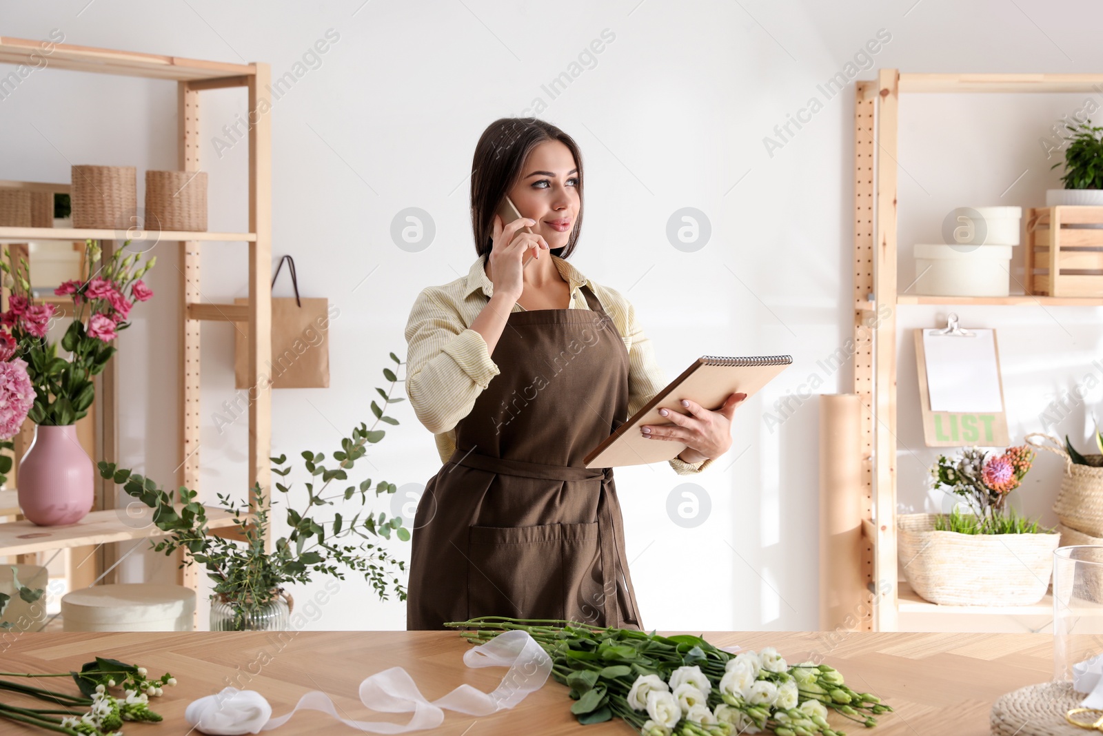 Photo of Florist with notebook talking on smartphone in workshop