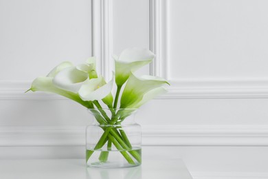 Photo of Beautiful calla lily flowers in glass vase on white table. Space for text