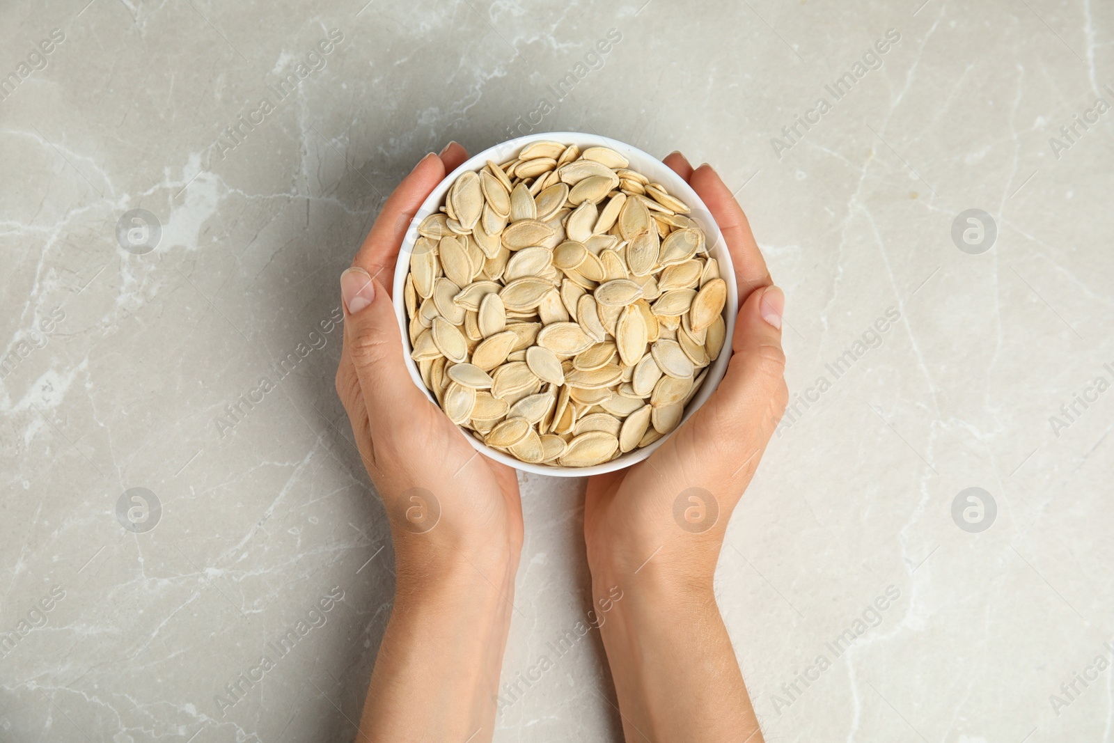 Photo of Young woman with bowl of raw pumpkin seeds at light grey marble table, top view