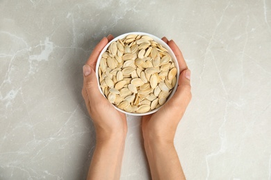 Photo of Young woman with bowl of raw pumpkin seeds at light grey marble table, top view