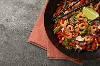 Shrimp stir fry with vegetables in wok and chopsticks on grey table, top view. Space for text