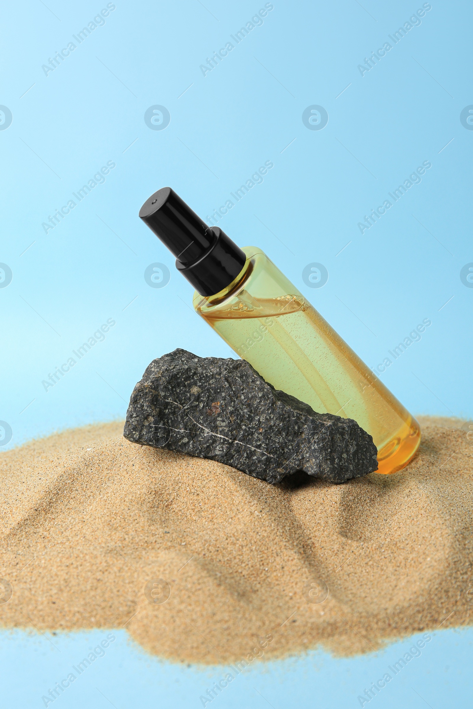 Photo of Bottle with serum and stone on sand against light blue background. Cosmetic product