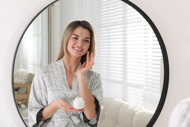 Photo of Beautiful young woman applying cream near mirror in room