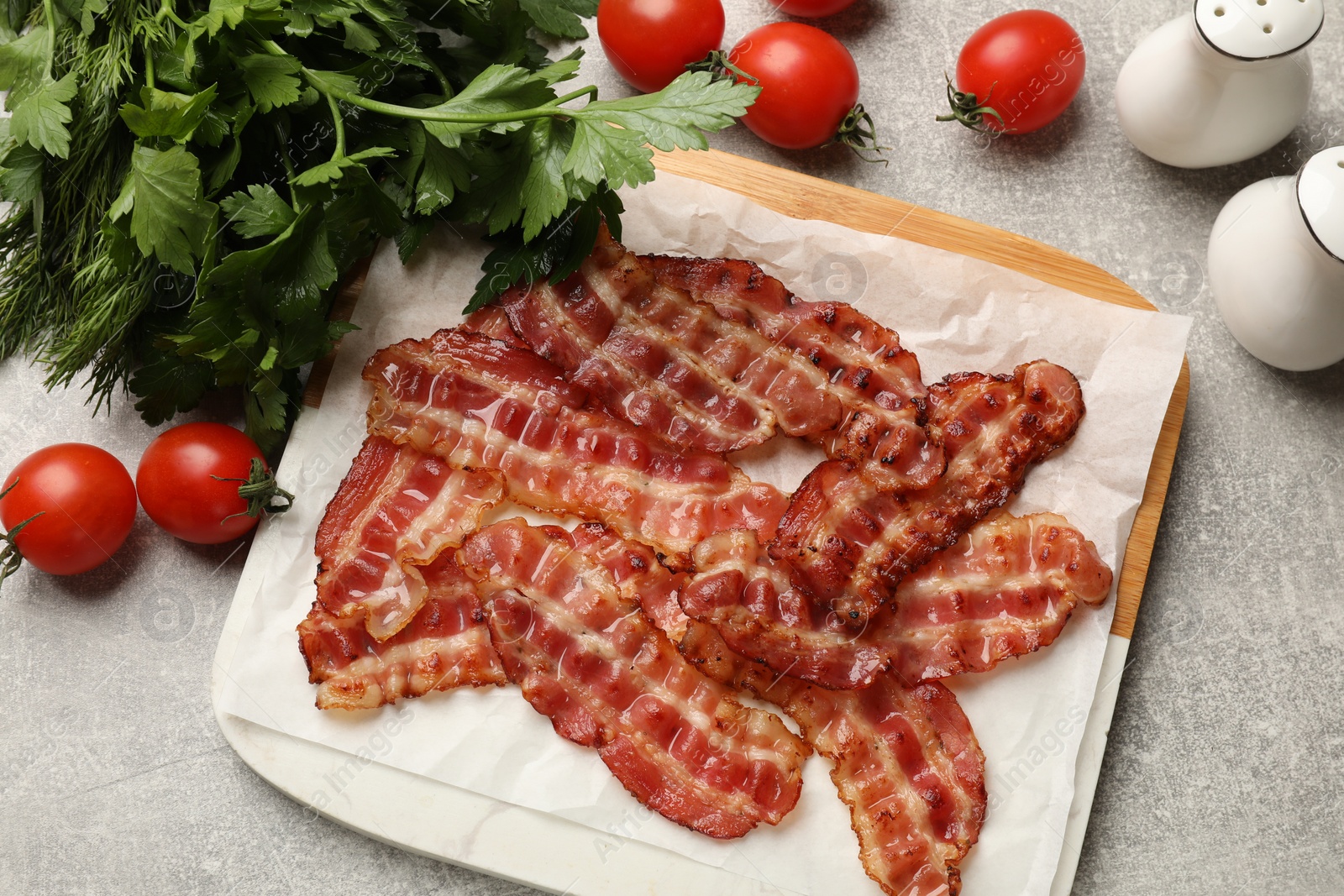 Photo of Fried bacon slices, tomato, parsley and spices on grey textured table, flat lay