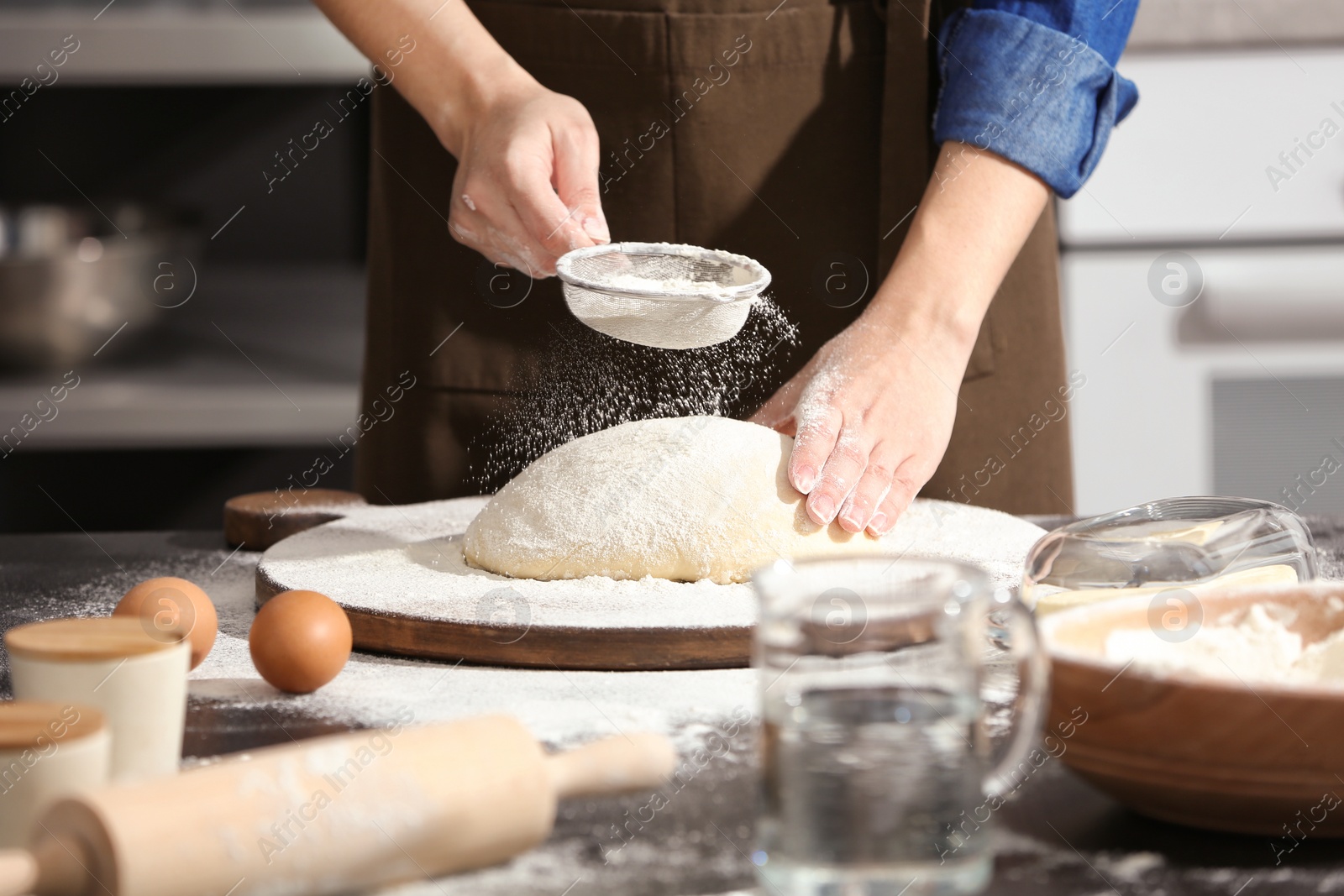 Photo of Woman sprinkling flour over dough on table in kitchen