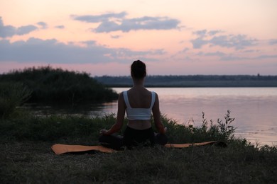Photo of Woman meditating near river in twilight, back view