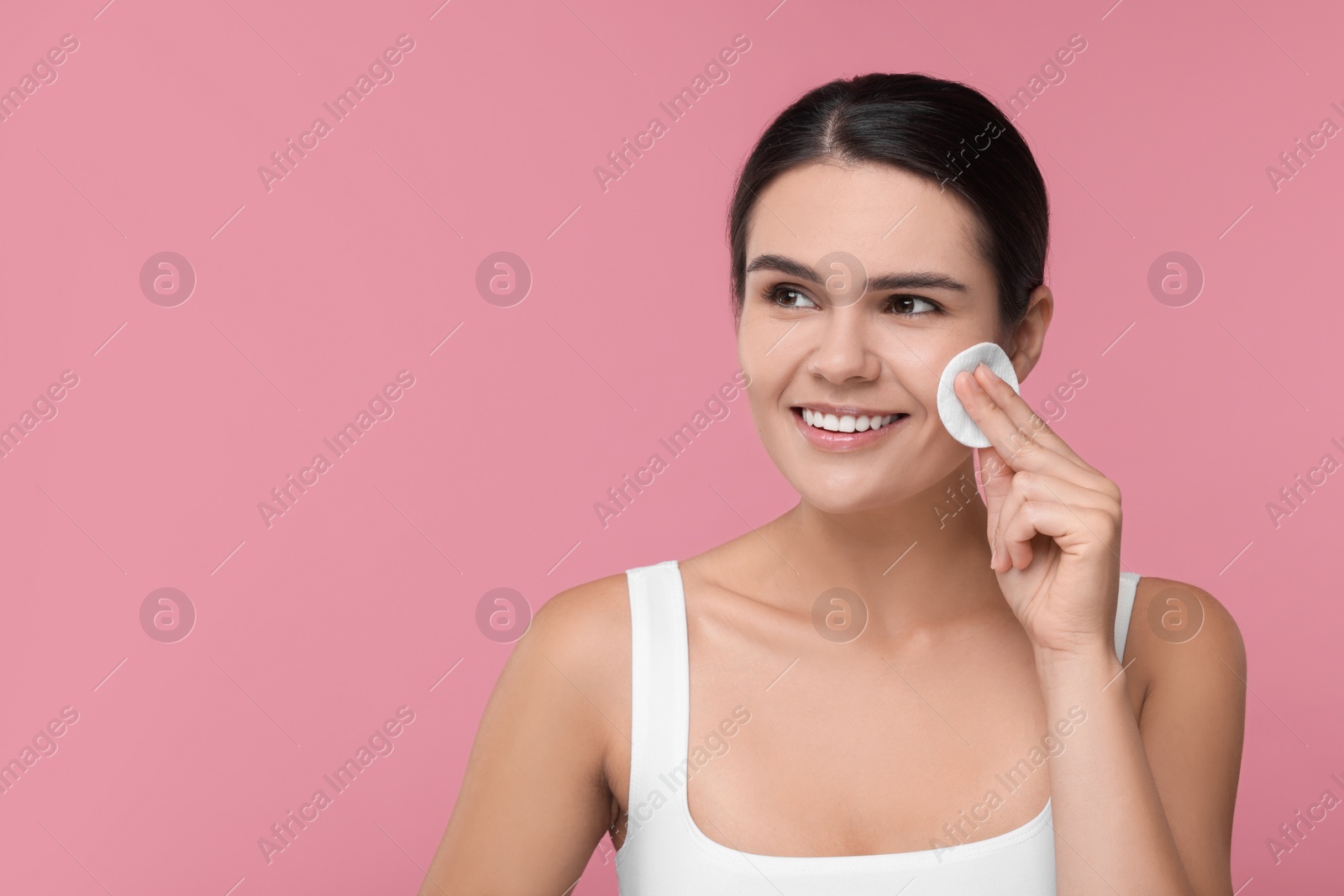 Photo of Young woman cleaning her face with cotton pad on pink background. Space for text