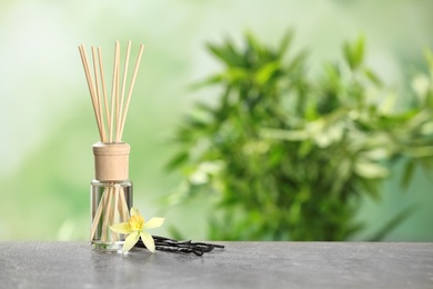 Reed air freshener with vanilla flower and sticks on grey table against blurred green background. Space for text