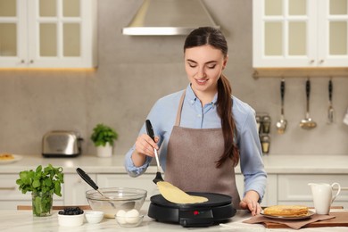 Photo of Happy woman cooking delicious crepe on electric maker at white marble table in kitchen