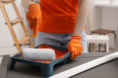 Photo of Woman taking orange paint with roller from tray at grey table indoors, closeup